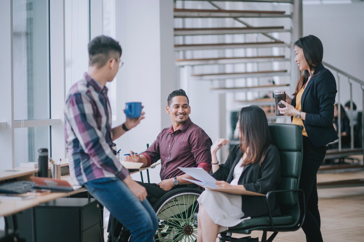 male worker in wheelchair having cheerful discussion leading conversation with colleague in creative office workstation